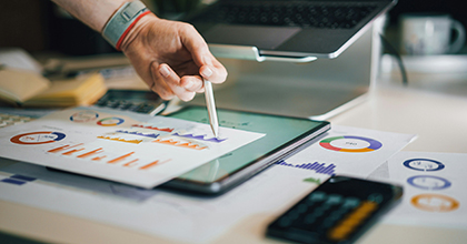 A person pointing a pen toward graphs and charts on a page laying on a table. 
