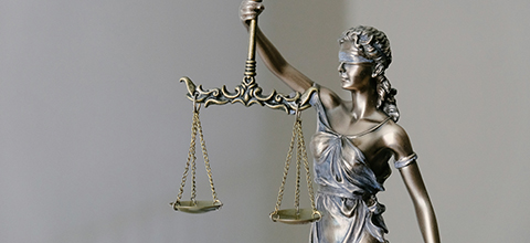 A metallic statue of Lady Justice holding balanced scales in her left hand, wearing a blindfold and draped gown, against a neutral background.
