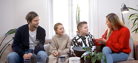 A husband and wife sit on a couch with their son and daughter having a conversation.