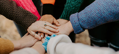 A group of people standing in a circle with their hands stacked together in the center, showing unity and teamwork. Several colorful sweaters and accessories are visible on their arms and hands.