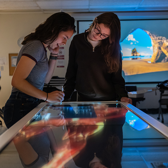 Two students stand over an illuminated interactive anatomy table, examining a digital 3D model together in a dimly lit classroom.
