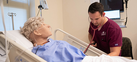 A nursing student in maroon scrubs uses a stethoscope to examine a medical training mannequin lying in a hospital bed, practicing a patient-care procedure in a clinical skills lab.