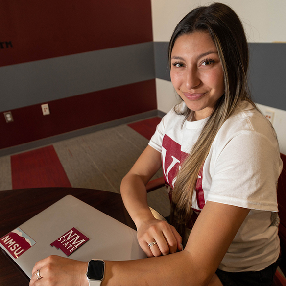 A female student smiles at the camera while sitting by her closed laptop. 